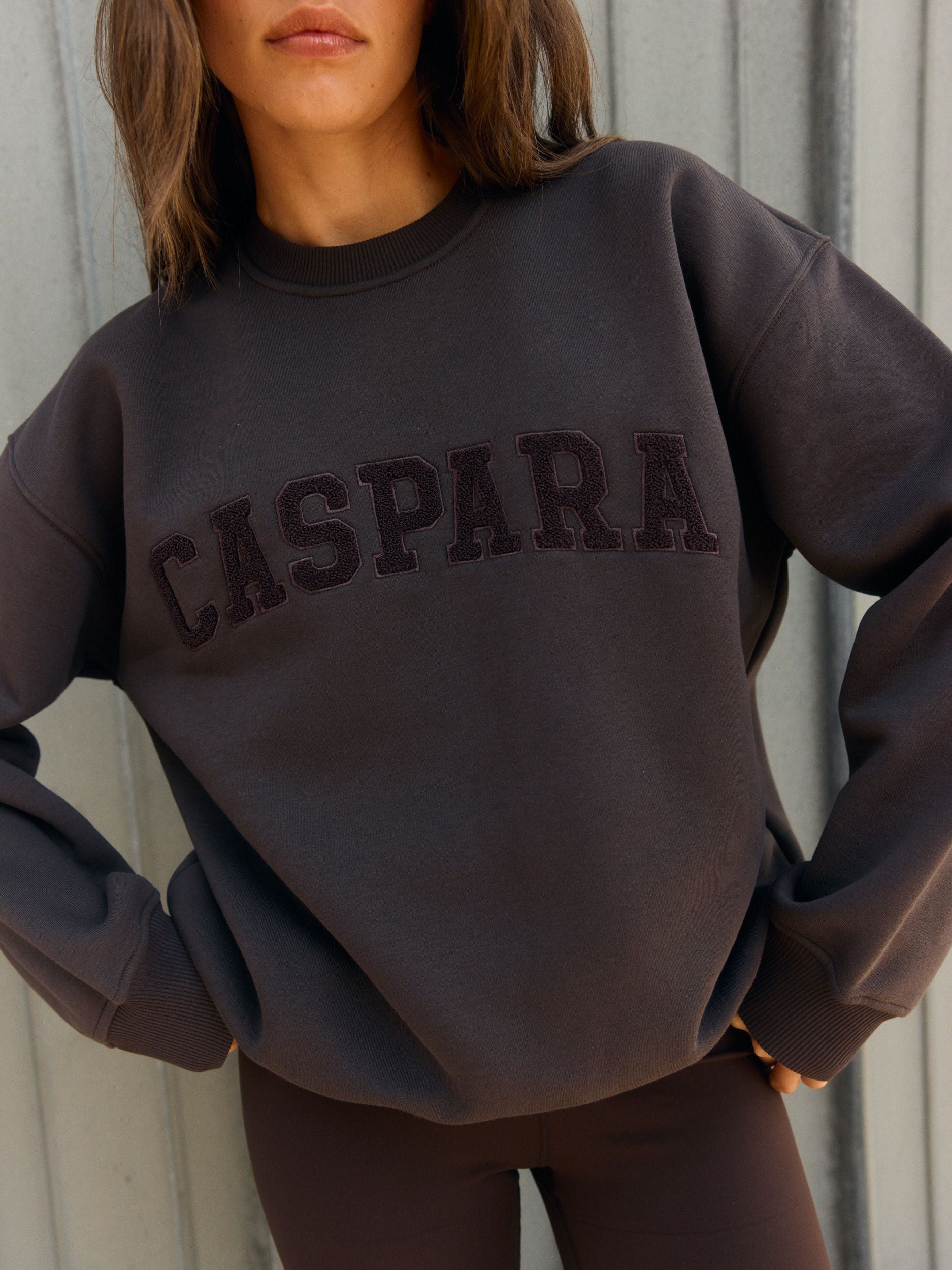 Woman wearing an espresso brown oversized crewneck sweatshirt with embroidered CASPARA lettering.